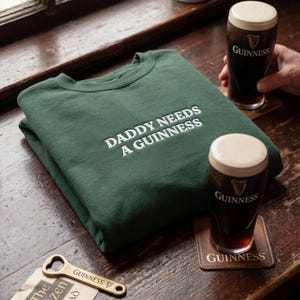 May include: A folded, forest green sweatshirt with the text "DADDY NEEDS A GUINNESS" in white. Two glasses of Guinness stout and a bottle opener are on a wooden surface.