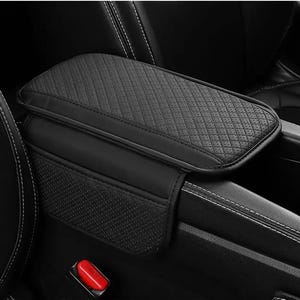 Puede incluir: Cubierta negra para reposabrazos de coche con una parte superior acolchada con diseño de diamantes. El reposabrazos está rodeado de cuero negro y tiene un pequeño bolsillo de almacenamiento. El interior del coche es negro con costuras blancas.