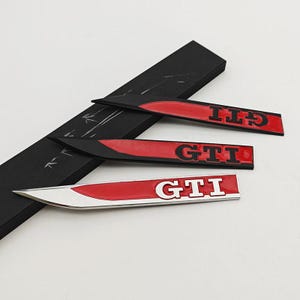 Pode incluir: Três emblemas de carro GTI. Os emblemas são vermelhos, pretos e prateados. O texto GTI é branco. Os emblemas estão sobre uma superfície preta.