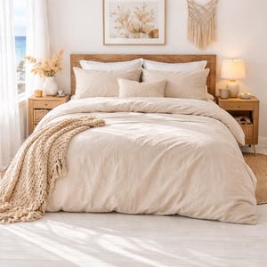 May include: A neutral-toned bedroom scene featuring a bed with a beige duvet cover and pillows. A woven throw blanket drapes over the bed. Wooden nightstands flank the bed, and a framed artwork hangs above the headboard. A lamp and decorative items complete the cozy aesthetic.