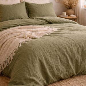 May include: A bed with olive green bedding, including a duvet cover and two pillowcases. A cream-colored fringed throw blanket is draped across the bed. The room has a neutral color palette with woven wall decor.