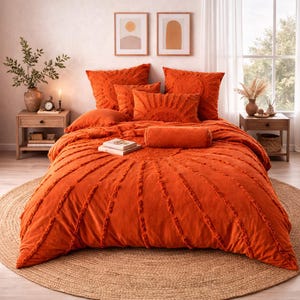 Op de afbeelding: Een oranje sprei met bijpassende kussens en een bolsterkussen. Het beddengoed heeft een getextureerd, getuft ontwerp. Het bed staat op een rond, geweven jute vloerkleed. Twee ingelijste kunstwerken hangen boven het bed.