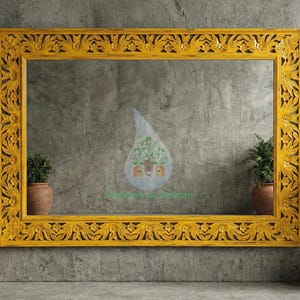 Op de afbeelding: Een rechthoekige spiegel met een felgele, ingewikkeld gesneden houten lijst. De lijst heeft een gedetailleerd bloemen- en bladerontwerp. De spiegel staat tegen een grijze muur met textuur, met potplanten aan weerszijden.