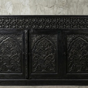 Puede incluir: Gabinete de madera negra con intrincados diseños florales tallados en las puertas y un patrón geométrico en la parte superior. El gabinete tiene tres puertas y está flanqueado por grandes macetas de terracota con plantas verdes.