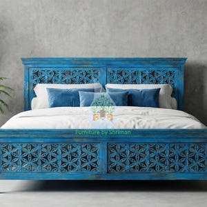 Puede incluir: Una cama de madera turquesa con intrincadas tallas florales. La cama está hecha con ropa de cama blanca y almohadas azules. A ambos lados hay grandes macetas de terracota con plantas. La habitación tiene una pared gris neutra.
