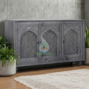 May include: A gray wooden cabinet with three doors and intricate carved detailing. The doors feature a lattice design, and the cabinet sits on short, rounded feet. The cabinet is placed against a concrete wall, with plants on either side.