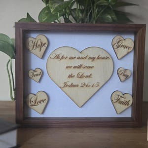 May include: Wooden framed wall art with a white background. The artwork features a large heart with the text "As for me and my house, we will serve the Lord. Joshua 24:15". Smaller heart-shaped cutouts spell out "Hope", "Trust", "Love", "Grace", "Pray", and "Faith".