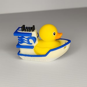May include: A yellow rubber duck in a white and blue boat. The duck has an orange beak and black eyes. The boat has blue wave designs and black smokestacks. A fun and whimsical bath toy.