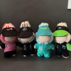 May include: Four plush dolls wearing racing team uniforms and visors. The dolls have bunny ears and are dressed in pink, black, teal, and green outfits. Each visor displays a team logo or number.