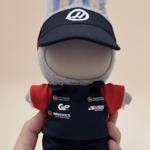 May include: A plush toy shaped like a rabbit wearing a black visor cap with a white logo and a navy blue racing uniform with red accents. The uniform has logos for "GP Barenice" and "awss". The toy has gray fur and blue inner ears.