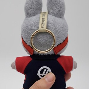 May include: A plush gray bunny keychain with a gold ring and red accents. The bunny wears a navy blue and red outfit with a white logo. The keychain has the text "STEIN MONSTER" on a beige strap.