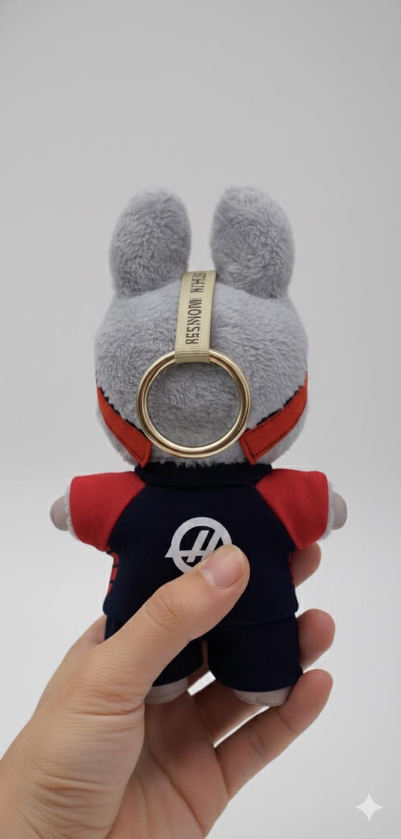 May include: A plush gray bunny keychain with a gold ring and red accents. The bunny wears a navy blue and red outfit with a white logo. The keychain has the text "STEIN MONSTER" on a beige strap.