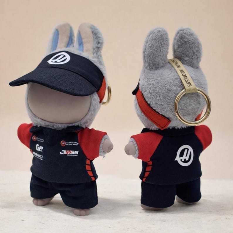 May include: A plush rabbit doll wearing a black visor with a white logo and a navy blue and red racing uniform. The doll has a gold keychain ring attached to its head. The uniform has the logos of sponsors.