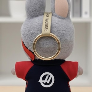 May include: A plush gray bunny toy wearing a navy blue and red outfit with a white logo. The bunny has a gold ring attached to its head with the text "RESIN MONSTER" on a beige strap.