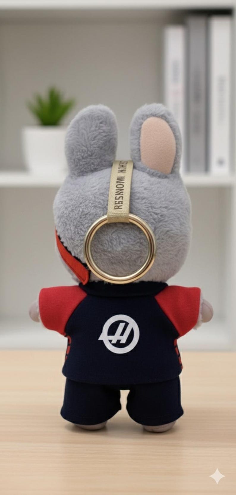 May include: A plush gray bunny toy wearing a navy blue and red outfit with a white logo. The bunny has a gold ring attached to its head with the text "RESIN MONSTER" on a beige strap.