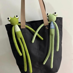 May include: A black tote bag with brown straps, adorned with two handmade crochet frog toys. The frogs are primarily green with white and blue accents, featuring large, expressive eyes and long limbs. The bag has a small owl logo.