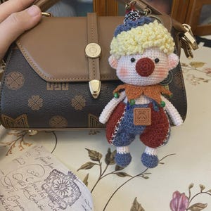 Könnte beinhalten: Eine braune und schwarze Handtasche mit goldenem Verschluss und Griff. Ein handgefertigter gehäkelter Clown-Schlüsselanhänger mit roter Nase, blau-rotem Outfit und gelben Haaren hängt an der Tasche. Ein Vintage-Dokument befindet sich im Vordergrund.