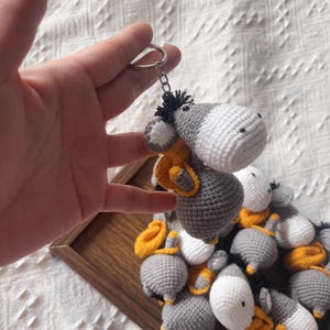 May include: Crocheted donkey keychain in gray, white, and yellow. The donkey has a white head, gray body, and a yellow scarf. The keychain is held by a hand, with several similar keychains in the background.