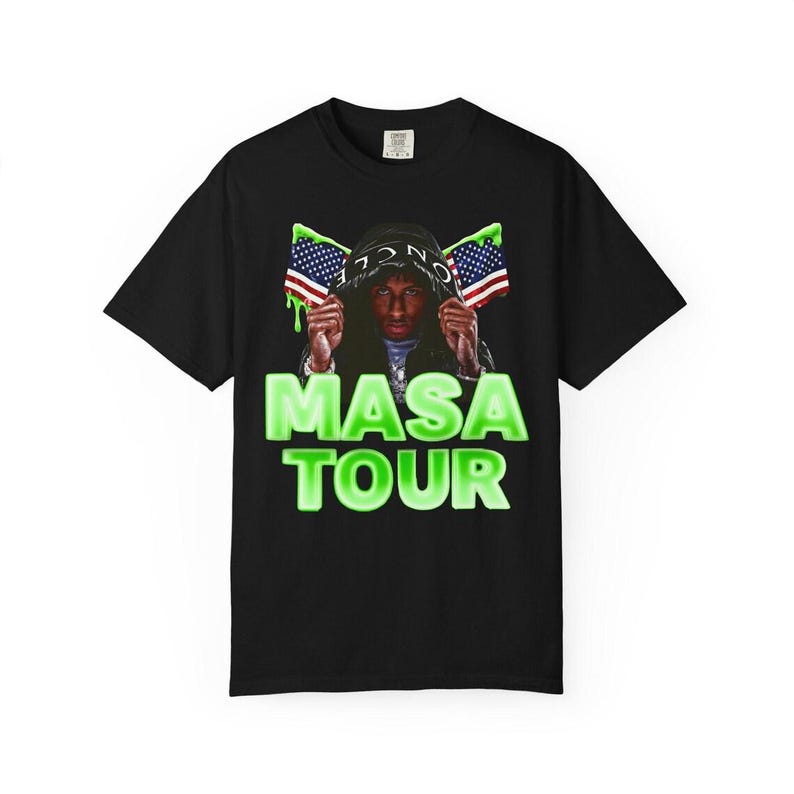 Pode incluir: Camiseta preta com um gr&aacute;fico de uma pessoa usando um capuz segurando bandeiras americanas. O texto "MASA TOUR" &eacute; exibido em verde neon abaixo.