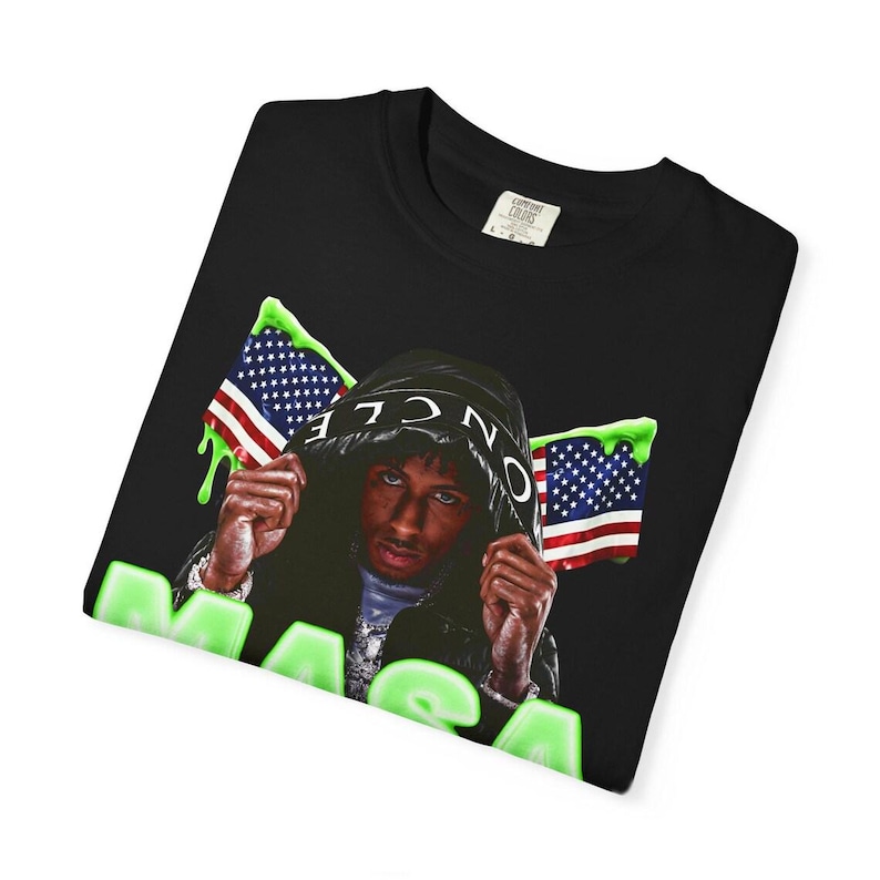 Pode incluir: Camiseta preta com um gr&aacute;fico de uma pessoa usando um casaco com capuz, bandeiras americanas e a palavra "MAGA" em verde. O design tem um efeito de gotejamento. A camisa est&aacute; dobrada.