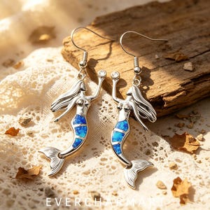 Può includere: Un paio di orecchini pendenti color argento con una silhouette di sirena, un inserto blu iridescente sulla coda e una piccola pietra trasparente in alto. Gli orecchini sono dotati di ganci e riportano la scritta EVERCHARM ART.