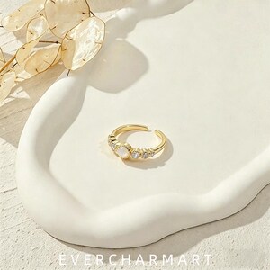 May include: A gold-toned open ring featuring a central round white stone flanked by two smaller clear stones on each side. The band has a smooth finish and an adjustable opening at the back. The ring is displayed on a white surface with dried leaves.