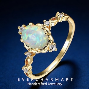 May include: A gold ring featuring an oval opal gemstone as the centerpiece, surrounded by smaller clear gemstones. The ring has a delicate, ornate design with the text "EVERCHARMART Handcrafted Jewellery" at the bottom.