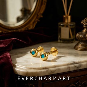 Könnte beinhalten: Zwei goldfarbene Labret-Stecker mit einem glatten, polierten kugelförmigen Kopf und einer kleinen, kreisförmigen, leuchtend türkisfarbenen Einlage in der Mitte. Die Schmuckstücke liegen auf einer Oberfläche, mit dem Text EVERCHARMART unten.