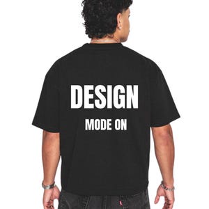 Op de afbeelding: Zwart T-shirt met de tekst "DESIGN MODE ON" in witte, vette letters. Het shirt heeft een klassieke ronde hals en lijkt gemaakt van een zacht materiaal. Het model draagt een donkere spijkerbroek.