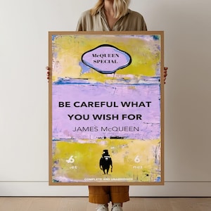 Puede incluir: Un cuadro enmarcado con un fondo abstracto amarillo, azul y morado. El texto "BE CAREFUL WHAT YOU WISH FOR" está centrado, con "JAMES MCQUEEN" debajo. Una silueta negra está en la parte inferior. El marco mide unos 15 cm de ancho.