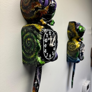 May include: Two whimsical cat-shaped wall clocks with a galaxy-themed design. The clocks feature a black clock face with white numbers and hands. The cat's head and body are painted with swirling colors of black, orange, green, and purple. The cat's eyes are purple.