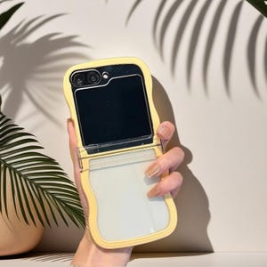 May include: A yellow phone case with a wavy design, open to reveal a black screen and a clear, rectangular section. The case has a camera cutout and a clear plastic hinge. The phone case is held in a hand.