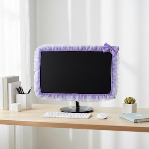 Pode incluir: Um monitor de computador com tela preta é emoldurado por uma capa de renda lavanda e um laço combinando. O monitor está em uma mesa de madeira com teclado, mouse e uma pequena suculenta.