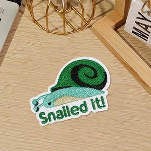 Peut inclure: Écusson brodé représentant un escargot de dessin animé avec une coquille verte et un corps bleu. Le patch porte le texte "Snailed It!" en vert. Le patch est sur une surface marron clair.