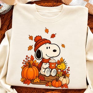 May include: A cream-colored crewneck sweatshirt featuring a graphic print of Snoopy and Woodstock. Snoopy wears an orange knit hat and scarf, holding a mug. They are surrounded by orange and yellow autumn leaves, two pumpkins, and berries.