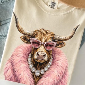 May include: A cream-colored t-shirt featuring a graphic of a brown cow wearing pink cat-eye glasses and a large, sparkling rhinestone necklace, surrounded by a fluffy pink faux fur collar. The shirt has a Comfort Colors brand label at the neck.
