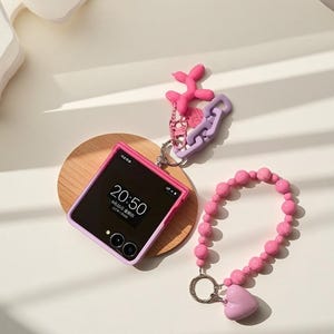 May include: A pink and purple smartphone showing 20:50 on its screen sits on a wooden coaster. A pink beaded bracelet with a heart charm and a keychain with a pink abstract shape and purple chain are also present.