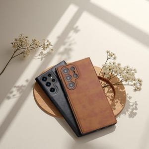 May include: Two phone cases, one black and one brown, are presented on a wooden coaster. The brown case has a textured, leather-like finish. The black case is smooth. Both cases feature multiple camera cutouts. White flowers and shadows enhance the presentation.