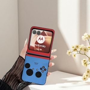 May include: A blue and red Motorola Razr 50 phone case designed to look like a retro video game controller. The case has a D-pad, buttons, and the Motorola logo. The phone is open, revealing the screen.