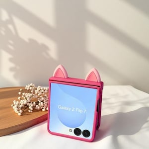 May include: A pink phone case with cat ears, designed for a Galaxy Z Flip 7. The case is a vibrant pink with a light blue screen. The phone is resting on a white surface with a wooden tray and white flowers in the background.