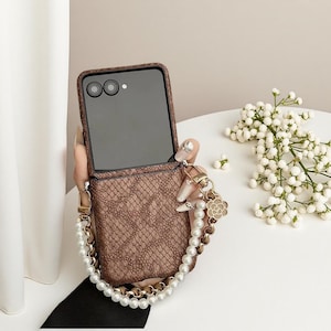 May include: A brown phone case with a snakeskin pattern, featuring a pearl and gold chain strap. The case has a decorative charm with a flower and a small gem. The phone screen is black.