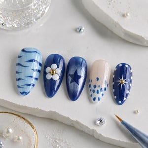 May include: Five painted artificial nails with various designs. The nails feature blue and white patterns, including stripes, a flower, a star, and polka dots. A fine-tipped brush and decorative elements are also visible.