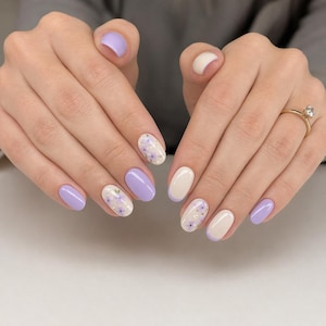 May include: Manicured nails in a variety of designs. Some nails are solid lavender, others are white with lavender floral patterns. A few nails are a cream color. A gold ring with a diamond is visible.