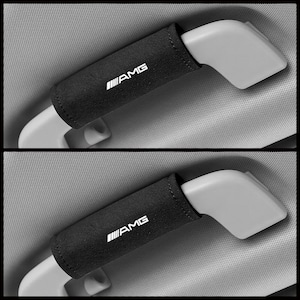 Puede incluir: Dos fundas negras para cinturón de seguridad con el texto blanco "AMG" impreso. Las fundas están envueltas alrededor de las manijas del cinturón de seguridad. El fondo es un interior de coche gris claro.