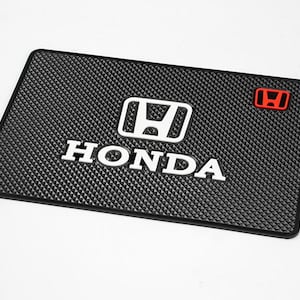 May include: Black rectangular mat with a textured surface and the Honda logo in white. The word "HONDA" is also in white, with a red Honda emblem in the upper right corner. The mat is likely for a car.