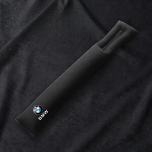 May include: A black seat belt cover with a BMW logo in white, blue, and black. The cover is rectangular and has a zipper at the top. The background is a dark, textured fabric.