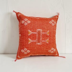 Peut inclure: Un coussin carré d'une teinte orange brûlé vibrante, orné de motifs géométriques brodés en blanc et jaune. Le coussin est agrémenté de petites franges délicates à chacun des quatre coins et présente une surface en tissu texturé.