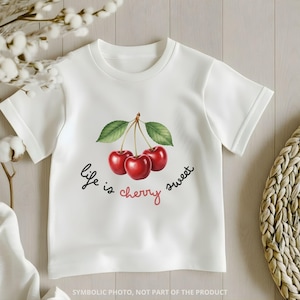 May include: White t-shirt featuring a watercolor illustration of three red cherries with green leaves. The text "life is cherry sweet" is written in black and red script. The shirt has short sleeves and a crew neck.