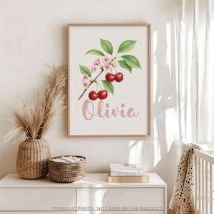 May include: A framed watercolor print featuring a cherry branch with red cherries, pink blossoms, and green leaves. The name "Olivia" is written in pink script below the branch. The frame is a light brown color.