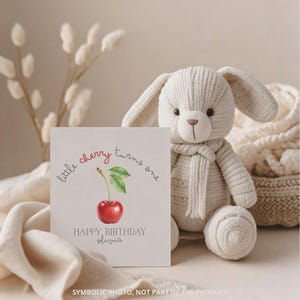 May include: A white knitted bunny plush toy with a cream-colored scarf sits next to a greeting card. The card features a watercolor illustration of a cherry and the text "little cherry turns one" and "HAPPY BIRTHDAY Olivia".